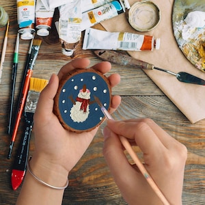 May include: A person is painting a snowman on a small, round wooden slice. The snowman has a red scarf and is set against a dark blue background with white dots. Paint tubes, brushes, and a palette are visible.