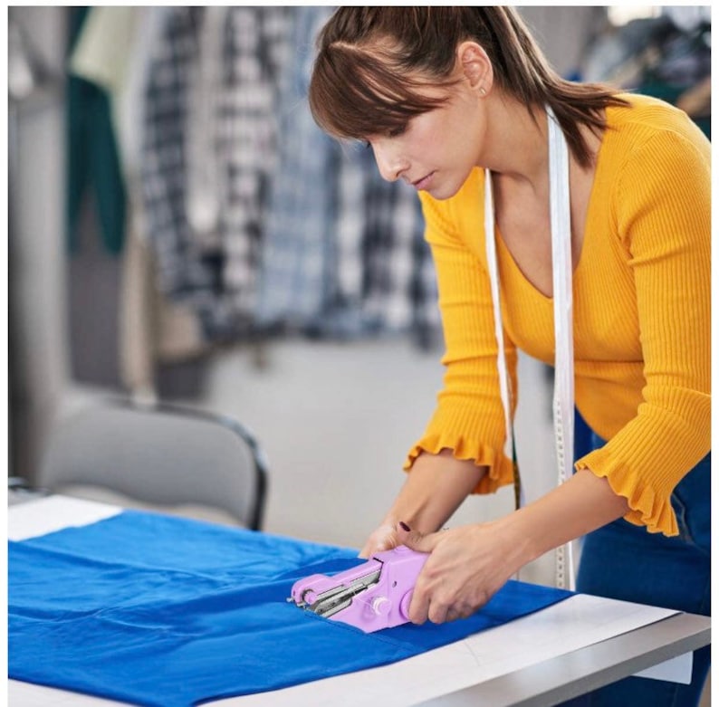 May include: A person using a pink handheld sewing machine to sew a blue fabric. The machine is battery operated and has a white cord.