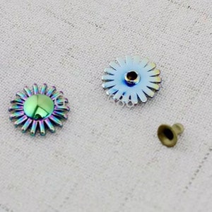 May include: Three metal embellishments in the shape of a flower with a center hole. The flower on the left is iridescent with a green center. The flower on the right is silver with a silver center. The small flower in the center is gold.