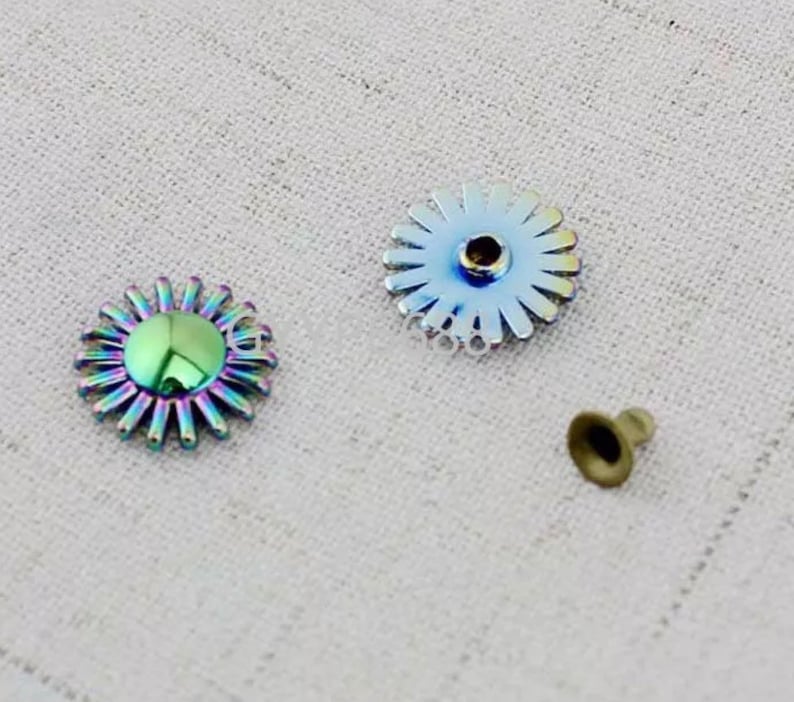 May include: Three metal embellishments in the shape of a flower with a center hole. The flower on the left is iridescent with a green center. The flower on the right is silver with a silver center. The small flower in the center is gold.