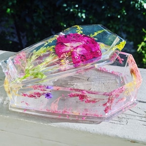 Op de afbeelding: Een transparante harskist in de vorm van een kist met een deksel. De kist is gevuld met geperste bloemen, waaronder een roze roos, en heeft een delicaat bloemenpatroon.