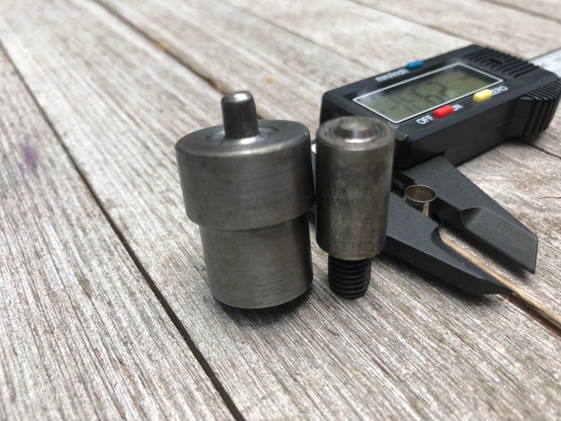 May include: Two metal tools for setting eyelets, one with a round head and the other with a pointed head, are measured with a digital caliper. The caliper reads 10.00 mm.