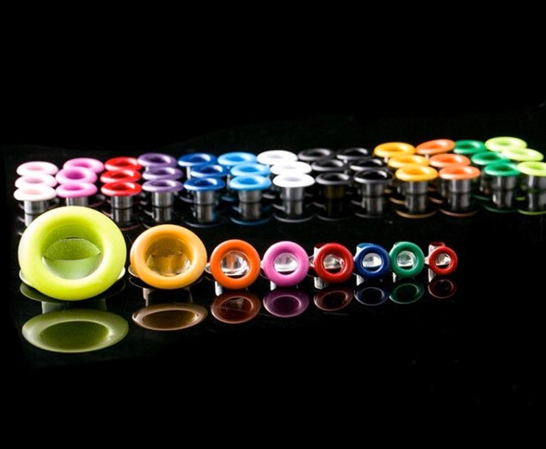 May include: Assortment of colorful metal eyelets in various sizes, including green, orange, red, blue, and purple. The eyelets are arranged in rows on a black surface.