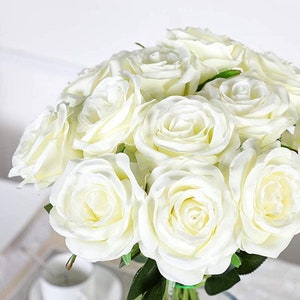 May include: A bouquet of white artificial roses arranged in a vase. The roses are made of a soft, silky material and have a realistic appearance.