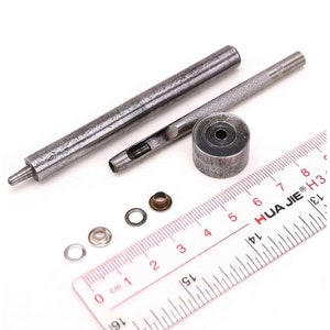 May include: A set of metal tools for installing eyelets, including a punch, a hammer, and a set of eyelets. The tools are laid out on a ruler with markings in centimeters.