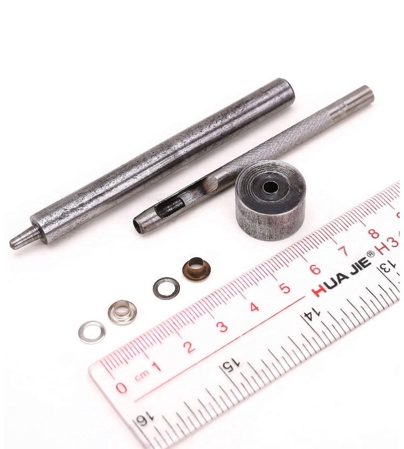 May include: A set of metal tools for installing eyelets, including a punch, a hammer, and a set of eyelets. The tools are laid out on a ruler with markings in centimeters.