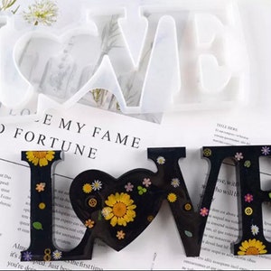 Puede incluir: Un letrero de resina negra con la palabra "LOVE" con detalles florales y engranajes. La "O" en forma de corazón está rellena con un gran girasol amarillo. Un molde de silicona transparente de la palabra "LOVE" está en el fondo. El letrero está sobre papel impreso.
