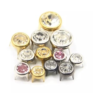 May include: Assortment of clear and pink rhinestone nailheads with gold and silver settings. The nailheads are arranged in a pyramid shape.