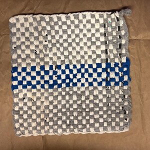 May include: Handmade square hot pad with a woven checkerboard pattern in gray and white, with a blue stripe across the center. The hot pad has a loop for hanging and is made of fabric.