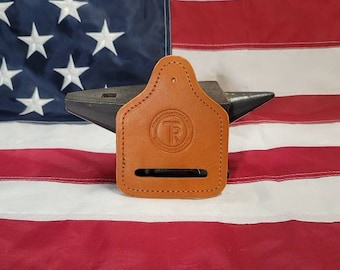 Op de afbeelding: Een bruine leren holster met een gestikte rand en een rond logo met de letters "TR" erin. De holster ligt op een aambeeld voor een Amerikaanse vlag.