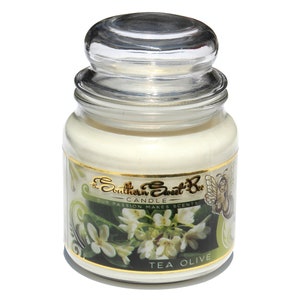 May include: A glass jar candle with a gold lid and label. The label features a green and white floral design with the text "The Southern Sweet Bee Candle Our Passion Makes Scents Tea Olive".