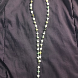 May include: A beaded necklace featuring round, light green and white stones strung on a silver chain. The necklace has a long, vertical design with a silver pendant at the bottom. The beads vary in color, with some appearing more gray or tan.