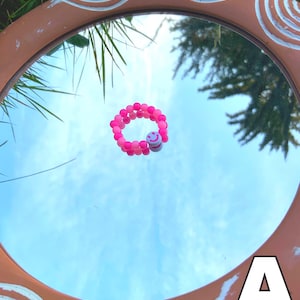 May include: A pink beaded bracelet with a white smiley face charm. The bracelet is reflected in a mirror.