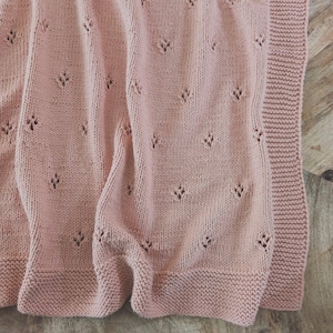 Op de afbeelding: Een zachtroze gebreide deken met een delicaat bladpatroon. De deken heeft een geribbelde rand en een getextureerd oppervlak. Het ontwerp is eenvoudig en elegant, geschikt voor een baby of als decoratief kleed.