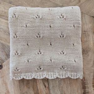 May include: A folded beige knit blanket featuring a repeating eyelet pattern arranged in vertical columns. The fabric has a smooth texture with a scalloped edge along the bottom border, showcasing a consistent stitch design throughout the piece.