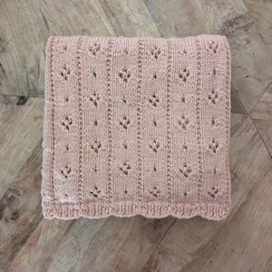 May include: A soft, pale pink knitted blanket featuring a repeating vertical pattern of delicate eyelet lace stitches. The fabric displays a textured, hand-crafted appearance with a consistent knit structure throughout the entire rectangular piece.