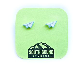 May include: A pair of white paper airplane earrings with blue accents on a green background. The earrings are displayed on a card with the text "South Sound Studios".