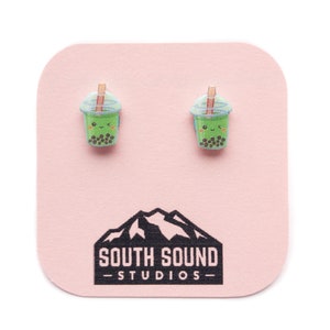 May include: A pair of green and white bubble tea earrings with a smiling face on each. The earrings are on a pink card with the text "South Sound Studios".