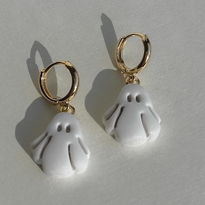 Pode incluir: Um par de brincos brancos em forma de fantasma com fechos de argola dourados. Os brincos têm um acabamento liso e brilhante e são desenhados com dois pequenos orifícios para os olhos e uma parte inferior curva.