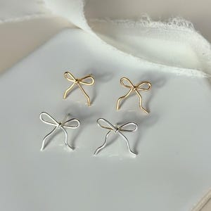 May include: Four bow-shaped earrings, two gold and two silver, are displayed on a white hexagonal tile. The earrings have a delicate, wire-like design. A white fabric is draped in the background, adding a soft texture to the image.