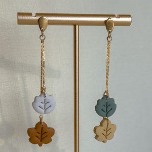May include: Dangling earrings with leaf-shaped charms in muted shades of brown, blue, green, and tan. The leaves hang from gold-coloured chains and posts, creating a delicate, nature-inspired accessory.