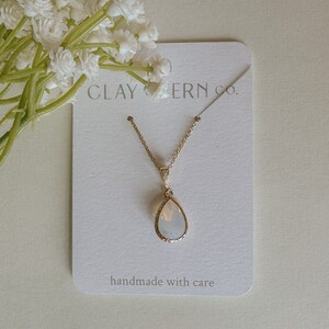 May include: A gold chain necklace with a teardrop-shaped white opal pendant. The pendant has a gold rim and is on a white card with the text "CLAY & ERN CO." and "handmade with care".