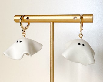 May include: A pair of white ghost-shaped earrings with black eyes hanging from gold hoops on a gold earring stand.