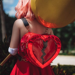 Może przedstawiać: Czerwony plecak w kształcie serca z przezroczystym panelem przednim i czarnym wzorem błyskawicy. Plecak nosi osoba z różowymi włosami i białą opaską na głowie.