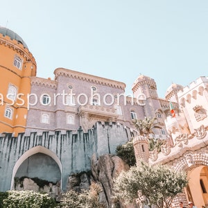 Op de afbeelding: Een uitzicht op het Pena National Palace in Sintra, Portugal. Het paleis is een mix van gele, witte en grijze steen met een verscheidenheid aan architectonische stijlen. Het paleis is omgeven door weelderige groene bomen en een stenen muur.