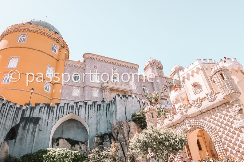 Op de afbeelding: Een uitzicht op het Pena National Palace in Sintra, Portugal. Het paleis is een mix van gele, witte en grijze steen met een verscheidenheid aan architectonische stijlen. Het paleis is omgeven door weelderige groene bomen en een stenen muur.