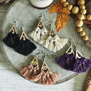 May include: Four pairs of Halloween-themed earrings with triangle-shaped wooden charms featuring different spooky designs. The earrings are made with macrame tassels in various colors, including black, white, purple, and brown.
