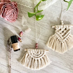 May include: Two beige macrame wall hangings with wooden dowels and fringe. The hangings are in the shape of a triangle and a half circle. A bottle of tea tree essential oil is in the foreground.