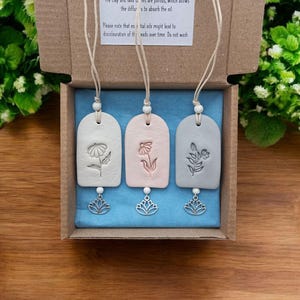 May include: Three clay essential oil diffusers in a box. Each diffuser is a different colour: white, pink, and grey. Each has a floral design and a silver lotus charm. The box includes instructions on how to use the diffusers.