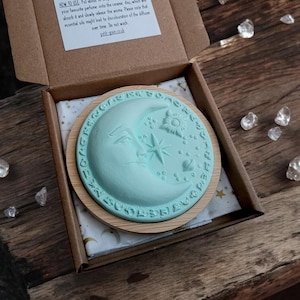 May include: A light blue ceramic essential oil diffuser with a moon and star design, in a wooden frame, inside a cardboard box. The box contains a printed instruction sheet. The diffuser is designed to release essential oil aromas. The measurements are in cm.