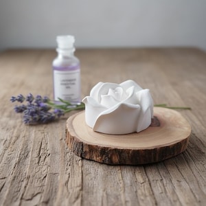 May include: A white rose-shaped ornament on a small, round wooden slice. A small bottle of lavender essential oil and a sprig of lavender are in the background. The items are arranged on a rustic wooden surface.