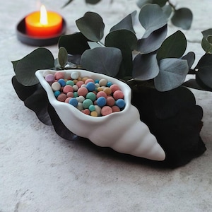 May include: A white ceramic seashell shaped dish filled with colourful glass beads. The dish is sitting on a light grey surface with green leaves in the background.
