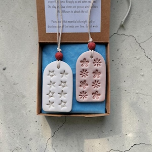 May include: Aromatherapy clay diffusers in a box. One is white with star cutouts, the other is pink with flower cutouts. Each has a red bead and a string for hanging. The box has text that reads "AROMATHERAPY CLAY & LAVA STONES DIFFUSERS".