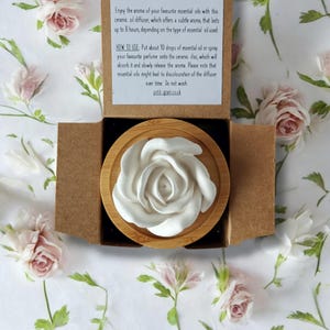 May include: A white ceramic rose diffuser sits on a wooden base. The diffuser is in a cardboard box with instructions on how to use it. "Enjoy the aroma of your favourite essential oils with this ceramic oil diffuser, which offers a subtle aroma, that lasts up to 8 hours, depending on the type of essential oil used. HOW TO USE: Put about 10 drops of essential oil or spray your favourite perfume onto the ceramic disc, which will absorb it and slowly release the aroma. Please note that essential oils might lead to discolouration of the diffuser over time. Do not wash petit-grain.co.uk"