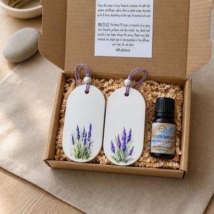 May include: A cardboard gift box containing two oval white ceramic diffusers with lavender illustrations, each with a purple hanging cord and bead, and a small amber glass bottle labelled Calm Mind pure essential oil blend with a black cap.