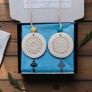 May include: Two white, circular clay diffusers with floral patterns, suspended by beige cords with yellow and turquoise beads. Each diffuser has a decorative metal charm. Presented in a black box with blue tissue paper.