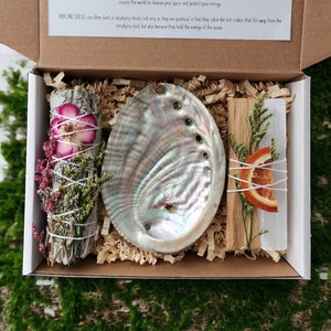 May include: A gift box set for smudging rituals, featuring a white sage smudge stick with dried flowers, a white selenite crystal, a piece of palo santo wood, and an abalone shell. The box is lined with brown paper.