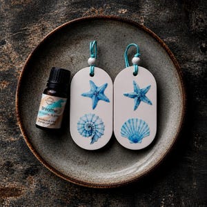May include: Two oval white ceramic hanging ornaments featuring blue starfish and seashell illustrations, each with a turquoise cord and white bead, alongside a small brown glass bottle labelled Breathe 100% Essential Oil Blend eucalyptus mint.
