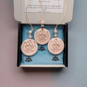 May include: Three pink agate diffusers with a lotus flower design and a metal lotus charm. Each diffuser hangs from a string with beads. The diffusers are displayed in an open box with text instructions.