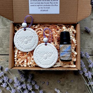 May include: A brown cardboard gift box containing two round white ceramic discs with embossed bee designs, each featuring a purple string and a small white bead. A small black bottle labelled Calm Mind essential oil blend is included.