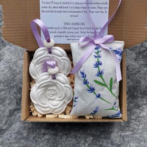 May include: A gift box containing two white ceramic flower-shaped oil diffusers and a lavender-scented fabric pouch. The box is lined with brown paper shreds and tied with a purple ribbon. The text on the box reads "Ceramic Oil Diffusers" and "French Lavender Bag".
