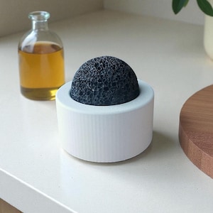May include: A black porous stone sits in a white cylindrical holder. A small glass bottle with amber liquid is in the background. The objects are on a white countertop, with a wooden board and greenery visible.