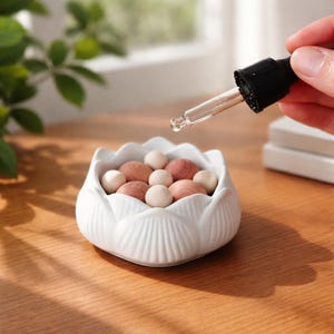 May include: A white ceramic lotus-shaped bowl filled with light brown and beige wooden balls. A dropper dispenses liquid above the bowl. The bowl sits on a wooden surface, suggesting a spa or aromatherapy theme.