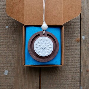 May include: A wooden pendant with a white snowflake design, suspended from a white cord, displayed in a brown cardboard box with blue tissue paper. The pendant features a dark brown outer ring and a white inner circle.