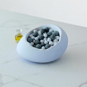 May include: A pale blue, egg-shaped bowl filled with grey and white spheres. A small, clear bottle with a yellow liquid sits to the left of the bowl. The objects are on a white marble surface.