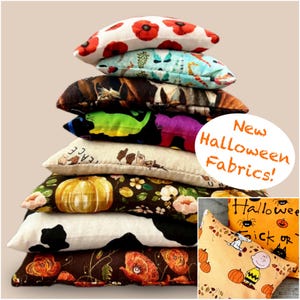May include: A stack of nine rectangular fabric pillows featuring various patterns including red poppies, teal ghosts, brown cats, neon cats, peace signs, pumpkins, cow spots, and floral prints. An inset shows orange fabric with Snoopy and text New Halloween Fabrics!
